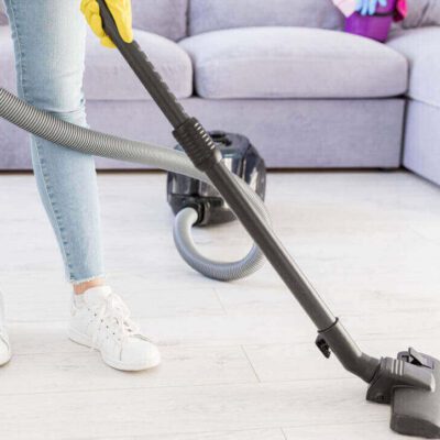 A low-angle shot shows a person's lower body as they vacuum a light-colored floor. They are wearing light blue jeans, white sneakers with white laces, and yellow gloves. The black hose and cleaning head of a canister vacuum cleaner are visible in the foreground, with the vacuum unit itself further back near a light grey sectional sofa.