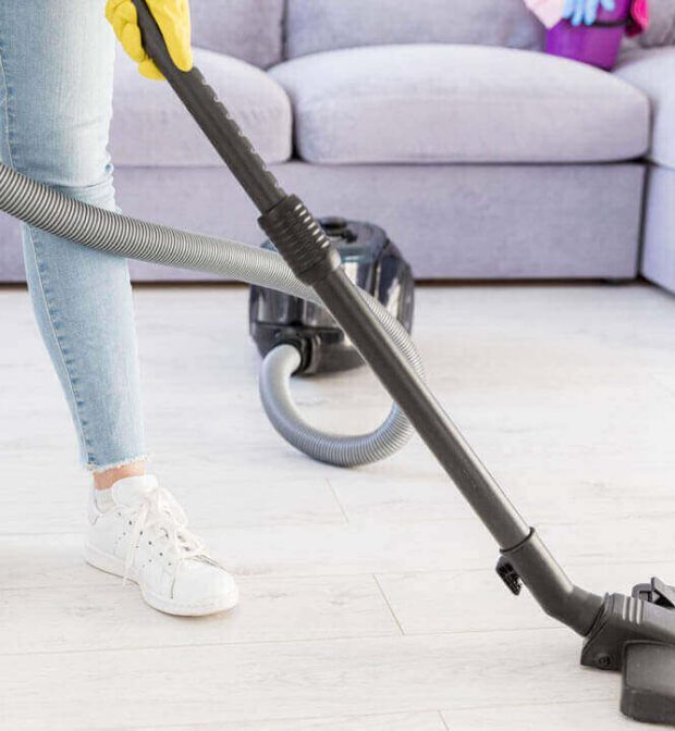 A low-angle shot shows a person's lower body as they vacuum a light-colored floor. They are wearing light blue jeans, white sneakers with white laces, and yellow gloves. The black hose and cleaning head of a canister vacuum cleaner are visible in the foreground, with the vacuum unit itself further back near a light grey sectional sofa.
