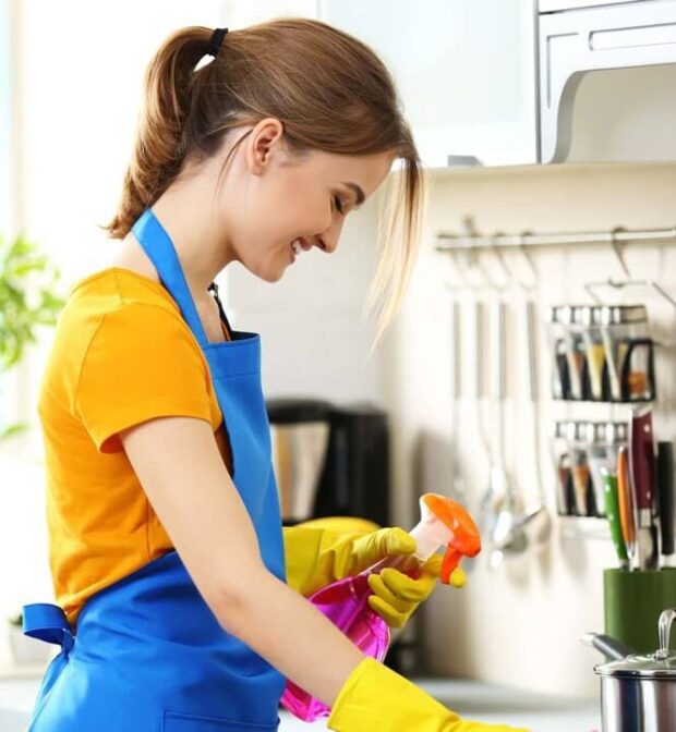 A smiling person with their hair tied back in a ponytail, wearing an orange t-shirt, a blue apron, and yellow rubber gloves, is shown from the side in a kitchen. They are holding an orange spray bottle in their right hand and appear to be cleaning a surface. In the background, there's a stove with pots, kitchen utensils hanging on a rack, and a green potted plant.